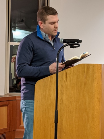 David Maloney: Image description - A man standing at a podium reading from a book.